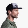 Un hombre de perfil, con una chaqueta verde, mira hacia la derecha mientras luce una elegante gorra Jockey Buff Trucker Sendel Black. La gorra muestra un diseño de árboles y montañas sobre un fondo gris claro.