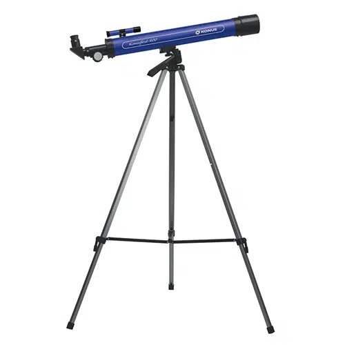 El refractor azul y negro Telescopio Konus Konusfirst 600 1723 está montado sobre un trípode plateado. El telescopio tiene un cuerpo cilíndrico largo con un ocular y un pequeño buscador acoplados cerca de la parte superior. El trípode completamente extendido tiene tres patas resistentes para sostener el telescopio.