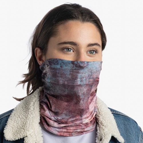 Una persona lleva una polaina para el cuello Bandana Buff Original EcoStrech Oxiron Multi que le cubre la nariz y la boca. Tiene el pelo castaño de longitud media y está vestida con una chaqueta azul con un cuello de borreguito blanco, sobre un fondo claro y liso.