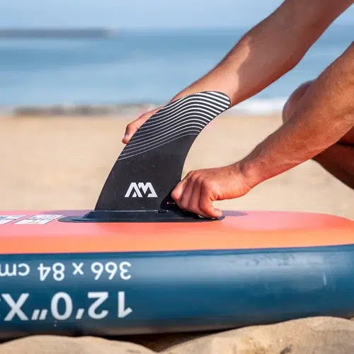 Una persona coloca una aleta Quilla Central Slide-In SUP Aqua Marina en una tabla de remo Aqua Marina roja y azul en la playa de arena, con el mar de fondo. La tabla muestra las marcas "12'0 X" y "366 x 84 cm".