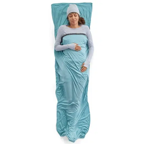 Una persona que lleva un gorro gris y una camiseta de manga larga está acostada boca arriba sobre un forro rectangular Sea To Summit Comfort Blend. El forro rectangular de color azul claro la envuelve por completo y se complementa con una funda de almohada a juego. Parece relajada y cómoda dentro de la combinación de confort de su configuración.