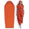Una mujer con un gorro y pantalones grises está envuelta en un saco de dormir naranja similar al Liner Sea To Summit Reactor Fleece Mummy Standard, famoso por su comodidad de estilo momia con forro polar. Junto a ella hay una vista desplegada que muestra su longitud total con capucha y laterales con cierre.