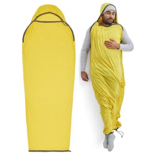 Una persona que lleva un saco de dormir amarillo portátil está de pie con una mano sobre el pecho. Junto a ella yace el estandarte de la momia del reactor Sea To Summit en su forma completa y extendida en el suelo. Lleva un gorro gris y calcetines.