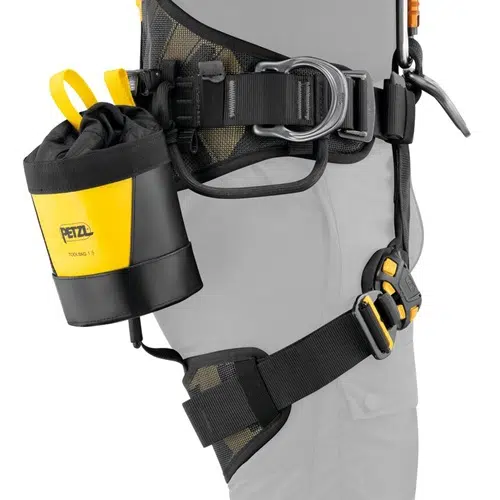 En primer plano, se ve un arnés de seguridad colocado alrededor de la cintura y las piernas, que muestra una Bolsa Portaherramientas Petzl Toolbag 1.5 en amarillo y negro sujeta al costado. El arnés está equipado con múltiples correas y hebillas de metal para garantizar una sujeción segura.