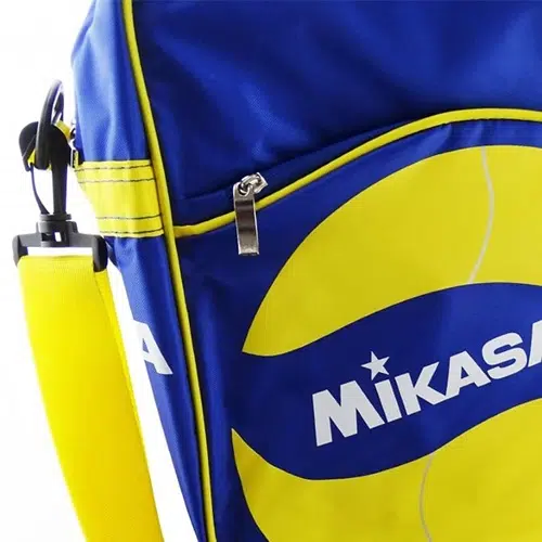 Primer plano del Bolso Portabalones Vóleibol MIKASA V200W, un bolso deportivo de color azul y amarillo que muestra su logotipo de forma destacada. El bolsillo frontal, parcialmente visible, incluye una cremallera y el bolso está equipado con una correa amarilla para el hombro para llevarlo cómodamente.