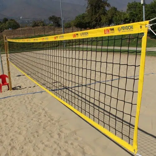 Una red de voleibol de color amarillo intenso se encuentra instalada sobre una cancha de arena rodeada de vegetación, lo que la convierte en un excelente lugar para un partido de vóleibol playa. Cerca de allí, se encuentra la silla Red Vóleibol Playa Uk Time Sport Profesional de 8,50 m x 1 m ubicada estratégicamente junto a la red. De fondo, se alzan majestuosas montañas bajo un cielo despejado, ofreciendo un entorno idílico para este deporte profesional.