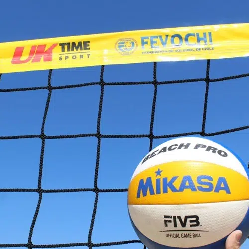 Una pelota de vóley playa Mikasa se coloca frente a una red contra un cielo azul claro. La red, una Red Vóleibol Playa Uk Time Sport Profesional de 8,50 m x 1 m, está adornada con los logotipos "UK TIME SPORT" y "FIVOCCHI", lo que le da un toque profesional a esta instalación de vóley playa.