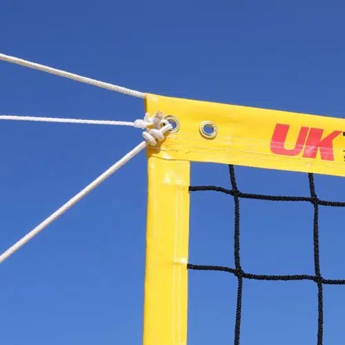 Primer plano de la esquina de una red de voleibol, contra un cielo azul claro, ideal para un partido profesional de voleibol de playa. La red tiene una cuerda blanca enhebrada a través de ojales de metal para mayor estabilidad y el texto parcial "UK" se ve en la banda roja superior.