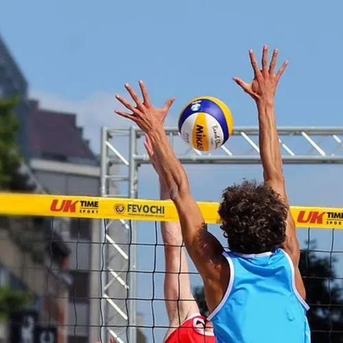 Dos jugadores profesionales de vóley playa compiten en la red. El jugador que lleva una camiseta azul salta con los brazos extendidos hacia arriba, cronometrando perfectamente su bloqueo. Mientras tanto, su oponente, vestido con un equipo Red Vóleibol Playa Uk Time Sport Profesional que mide 8,50 m x 1 m, es visible detrás de la red. El balón de voleibol flota sobre ellos en este vívido escenario al aire libre del Reino Unido.
