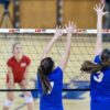 Durante un emocionante partido de voleibol en sala, dos jugadores con camisetas azules saltan con los brazos extendidos para bloquear la pelota en la red. Mientras tanto, un jugador oponente que viste la camiseta roja de voleibol Uk Time Sport Indoor de 9,50 m x 1 m se prepara para devolverla al otro lado de la cancha.