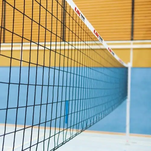 Vista en primer plano de una red de voleibol ubicada dentro de una cancha cubierta. La red, con la marca Red Vóleibol UK Time Sport Indoor de 9,50 m x 1 m, tiene una cinta superior blanca y el fondo presenta una pared azul con líneas horizontales amarillas y blancas encima. El piso de la cancha es parcialmente visible en la parte inferior.