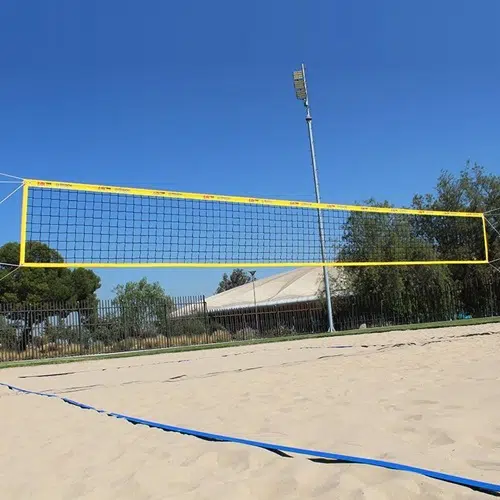 Una cancha de voleibol de playa con una red roja está instalada en una zona de arena. Las líneas azules delimitan los bordes de la cancha. En el fondo, hay árboles, una cerca, un poste de luz y un cielo azul claro, perfecto para un juego profesional con la Red Vóleibol Playa Uk Time Sport Profesional 8.50m x 1m.
