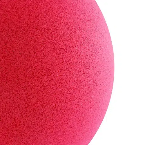 Un primer plano de una esponja de maquillaje de color rosa vibrante llena el lado izquierdo de la imagen, similar a un Balón de Esponja Uk Time Sport de 6″. Su textura porosa, forma redonda y superficie suave se destacan sobre un fondo blanco, lo que resalta su color intenso.