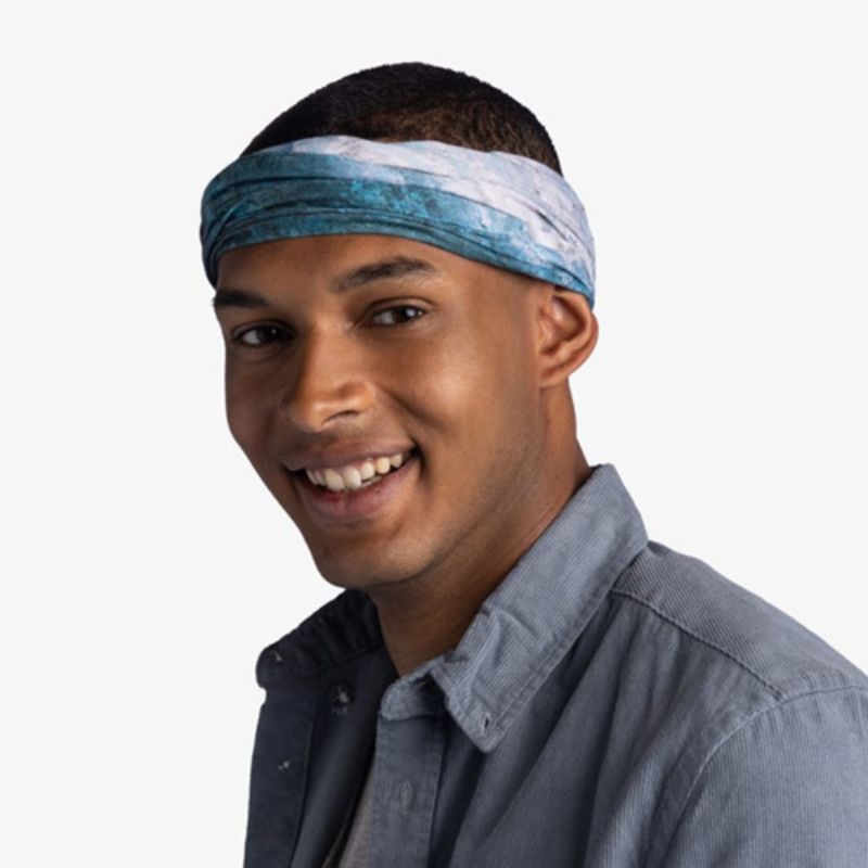 Una persona sonríe mientras usa el Bandana Buff CoolNet UV Watsea Blue Surfrider y una camisa gris abotonada, de pie sobre un fondo blanco liso.