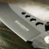 Primer plano de una Navaja Boker Magnum US Navy Seals 01MB856 con la hoja grabada "MAGNUM by BÖKER". La hoja, que recuerda a las preferidas de los SEAL de la Marina de los EE. UU., presenta cortes circulares cerca del mango y reposa sobre una superficie metálica.