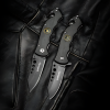 Dos navajas plegables, similares a la Navaja Boker Magnum US Navy Seals 01MB856, presentan un diseño negro con hojas parcialmente dentadas y mangos texturizados acentuados por detalles de emblemas redondos. Colocadas sobre una superficie de cuero, muestran hojas con acabado mate con grabados prominentes de la marca.