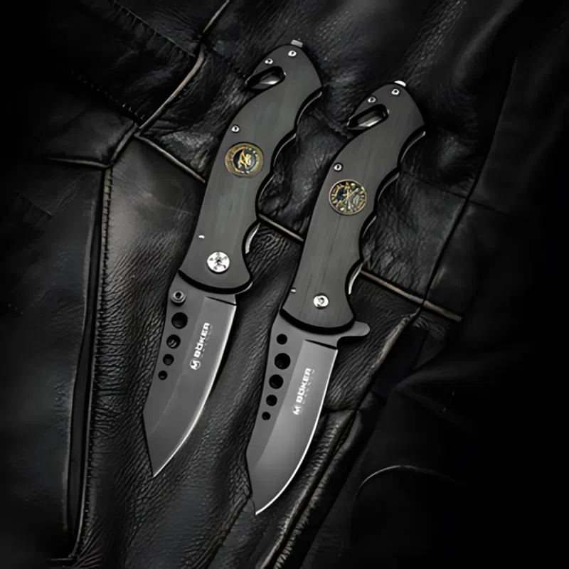 Dos navajas plegables, similares a la Navaja Boker Magnum US Navy Seals 01MB856, presentan un diseño negro con hojas parcialmente dentadas y mangos texturizados acentuados por detalles de emblemas redondos. Colocadas sobre una superficie de cuero, muestran hojas con acabado mate con grabados prominentes de la marca.