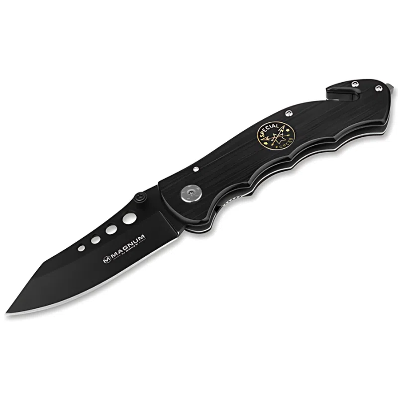 Navaja de bolsillo plegable negra con hoja parcialmente dentada. El mango presenta un botón plateado y un emblema circular con elementos náuticos. Las palabras "Navaja Boker Magnum Us Navy Seals 01MB856" están impresas en la hoja y el mango, respectivamente.