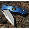 Una navaja plegable azul Boker Magnum Law Enforcement 01MB365, con una hoja plateada y un emblema redondo dorado en el mango, está abierta sobre una superficie rocosa. La hoja, con la inscripción "MAGNUM", brilla a la luz del sol.