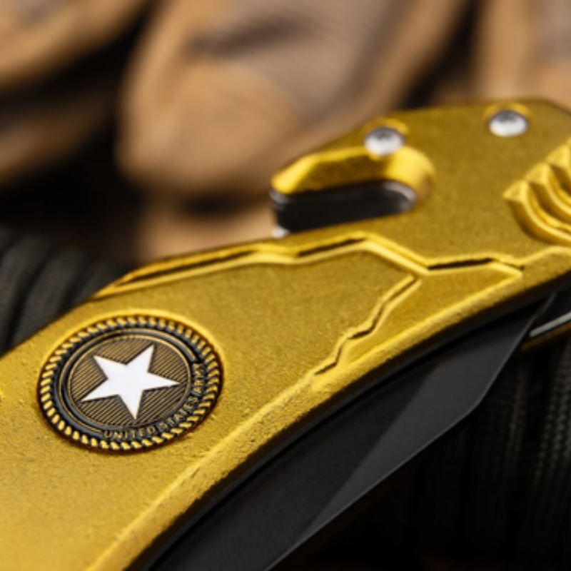 Una imagen en primer plano del mango de la navaja de bolsillo Navaja Magnum Army Rescue 01LL471 muestra su acabado dorado con un emblema de estrella texturizado. En el fondo, un par de guantes color canela complementan la escena, resaltando el diseño elegante y los detalles intrincados de la navaja.
