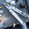 Una multiherramienta Leatherman Curl 832932, con los alicates extendidos, descansa sobre una superficie en medio de una variedad de coloridas moscas de pesca y pequeñas botellas, evocando un animado arreglo o preparación de pesca.
