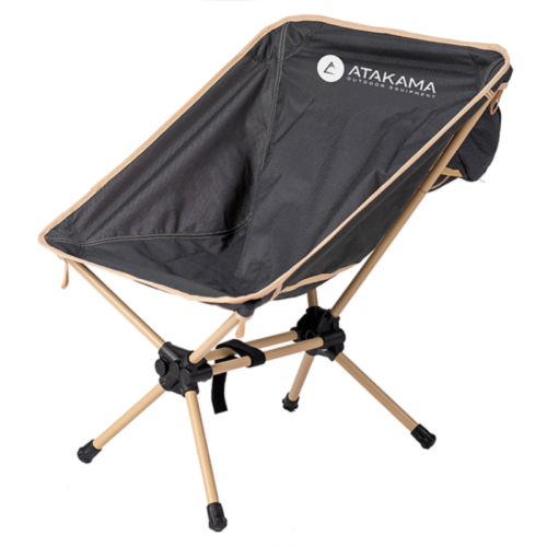 Silla de camping portátil con asiento de tela negra y armazón beige. El asiento tiene el logotipo de "Silla Camping Atakama Outdoor Láscar" y el texto "Outdoor Equipment" impreso en el respaldo. La silla tiene cuatro patas con juntas y tapas de plástico negro.