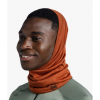 Una persona lleva una polaina para el cuello Bandana Buff Merino Lightweight Solid en color naranja, que le cubre la cabeza y el cuello con un logotipo en la tela, combinada con una chaqueta ligera de color verde claro. Sonríe y mira ligeramente hacia la derecha sobre un fondo blanco liso.