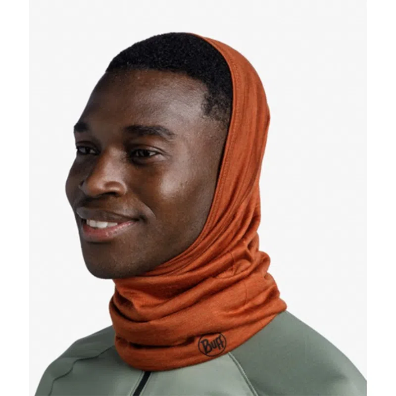 Una persona lleva una polaina para el cuello Bandana Buff Merino Lightweight Solid en color naranja, que le cubre la cabeza y el cuello con un logotipo en la tela, combinada con una chaqueta ligera de color verde claro. Sonríe y mira ligeramente hacia la derecha sobre un fondo blanco liso.
