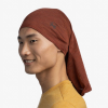 Una persona que lleva una Bandana Buff Merino Lightweight Multistripes en color naranja quemado, sonriendo y mirando ligeramente hacia la derecha. Está vestida con una camiseta amarilla con múltiples rayas sobre un fondo blanco liso.