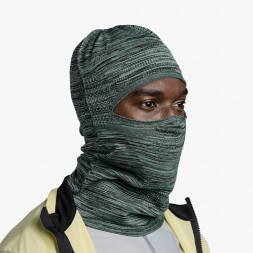 La persona, que luce el Pasamontañas Buff DryFlx Pro Solid Jade en un diseño verde texturizado que cubre la mayor parte de su rostro y cabeza, también está vestida con una chaqueta beige y negra sobre una parte superior gris claro, todo sobre un fondo blanco liso.