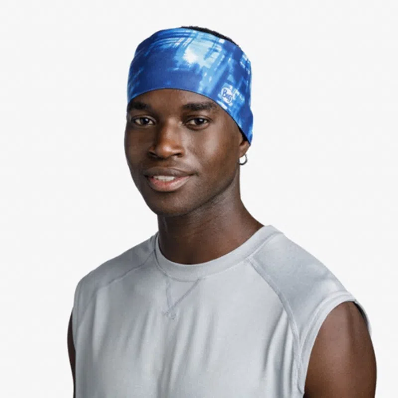 Una persona sonríe a la cámara mientras usa una diadema Cinta Buff CoolNet UV Wide Attel Blue y una camisa sin mangas gris claro, sobre un fondo blanco liso.