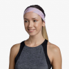 Una mujer con el pelo largo recogido en una cola de caballo luce una cinta para el pelo Cinta Buff CoolNet UV Slim Shane Orchid de color pastel y una camiseta gris y negra sin mangas. Sonríe sobre un fondo blanco liso, encarnando sin esfuerzo el encanto del estilo Buff CoolNet UV.