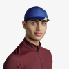 Una persona que viste una gorra Jockey Buff Pack Cycle Akim Cobalt y una camisa con cremallera color burdeos mira a la cámara sobre un fondo blanco liso.