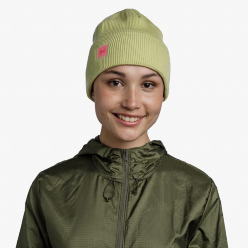 Una persona sonríe, vistiendo un gorro Buff CrossKnit Solid Sheen Yellow verde claro con un logotipo cuadrado rosa combinado con una chaqueta verde oliva, sobre un fondo blanco liso.