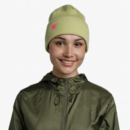 Una persona sonríe, vistiendo un gorro Buff CrossKnit Solid Sheen Yellow verde claro con un logotipo cuadrado rosa combinado con una chaqueta verde oliva, sobre un fondo blanco liso.