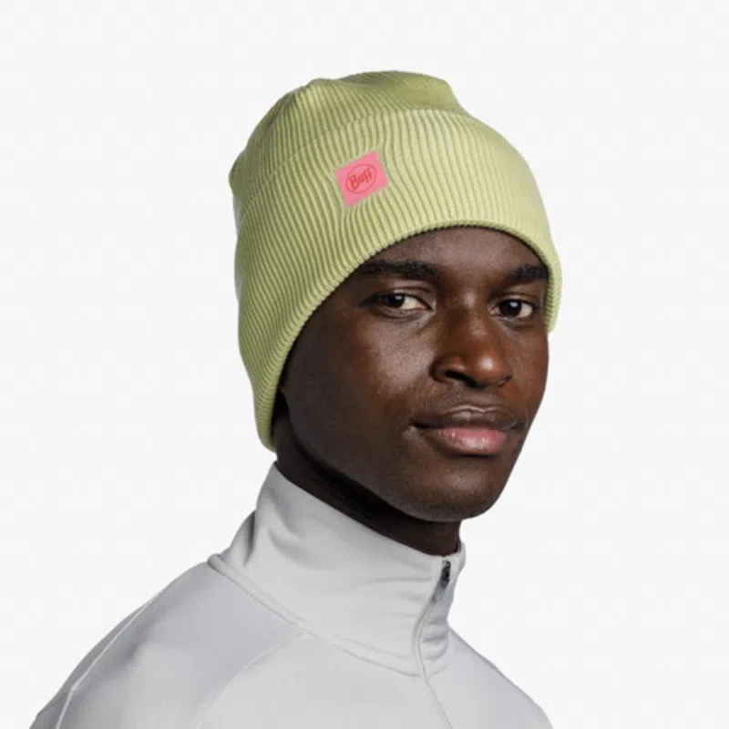 Una persona que lleva un gorro amarillo con un logotipo rosa, similar al estilo Gorro Buff CrossKnit Solid Sheen, lo combina con una camiseta con cierre de cremallera gris claro. Mira de reojo contra un fondo blanco liso.