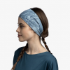 Una mujer con cabello castaño largo peinado en dos trenzas lleva una diadema Cinta Buff CoolNet UV Wide Laven Mist. Su camiseta azul complementa la diadema, mientras mira hacia la izquierda sobre un fondo blanco liso.