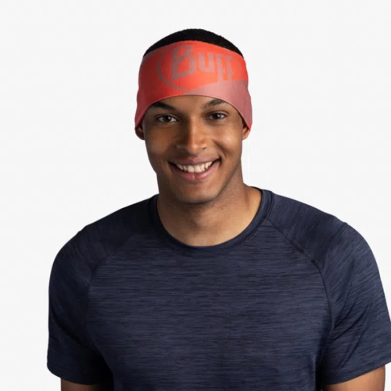 Una persona sonríe mientras usa una diadema Cinta Buff CoolNet UV Wide Arthy Nectarine con un logotipo y una camiseta azul marino sobre un fondo blanco liso.
