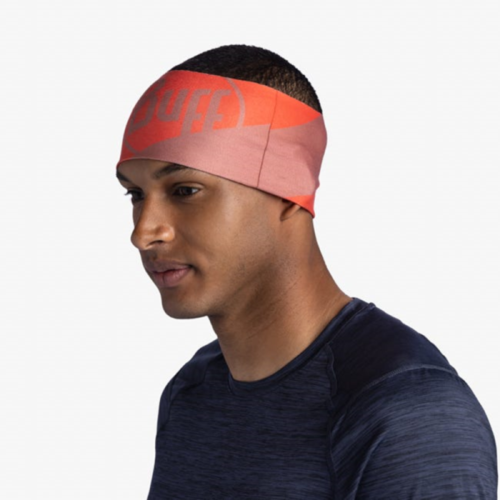 Una persona que lleva una diadema Cinta Buff CoolNet UV Wide Arthy Nectarine rosa y roja con la palabra "BUFF" se encuentra sobre un fondo liso, vestida con una camisa azul oscuro.