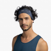 Una persona con cabello negro ondulado viste una diadema Cinta Buff Fastwick Bonsy Graphite y una camisa azul oscuro sin mangas, de pie sobre un fondo gris claro.