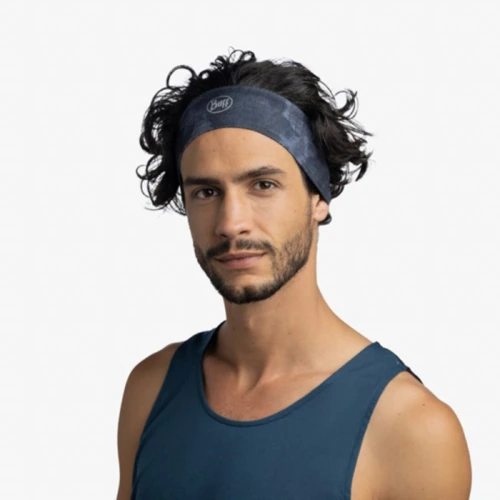 Una persona con cabello negro ondulado viste una diadema Cinta Buff Fastwick Bonsy Graphite y una camisa azul oscuro sin mangas, de pie sobre un fondo gris claro.