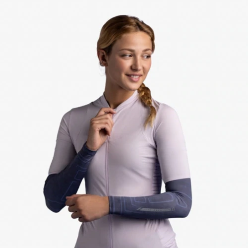 Una persona que viste una camiseta de ciclismo violeta claro y mangas de brazo Buff está de pie sobre un fondo liso, mirando hacia su derecha con una leve sonrisa. Lleva el pelo recogido en una trenza sobre un hombro.