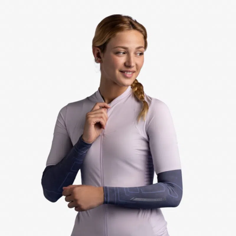 Una persona que viste una camiseta de ciclismo violeta claro y mangas de brazo Buff está de pie sobre un fondo liso, mirando hacia su derecha con una leve sonrisa. Lleva el pelo recogido en una trenza sobre un hombro.