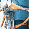Un primer plano de una persona que lleva el arnés Arnés Petzl Sitta 2024 en un paisaje nevado. El arnés presenta un diseño naranja dominante con elegantes detalles en blanco y está equipado con mosquetones y el equipo de escalada esencial, combinando a la perfección estilo y seguridad para escaladores apasionados.