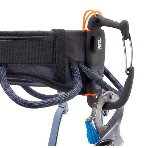 Primer plano de un equipo de escalada que muestra un arnés Arnés Petzl Corax 2024 equipado con un mosquetón, bloqueador y presillas de cuerda adjuntas. Los componentes están diseñados para garantizar la seguridad y la eficiencia en la escalada. El equipo parece resistente y bien construido.