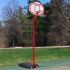 Un aro de baloncesto rojo Uk Time Sport con pedestal Zy-003 se encuentra en una posición destacada en una cancha al aire libre, rodeado de áreas verdes y árboles. Este aro de baloncesto portátil se recorta contra un cielo despejado, creando un entorno sereno y natural para los amantes del deporte.