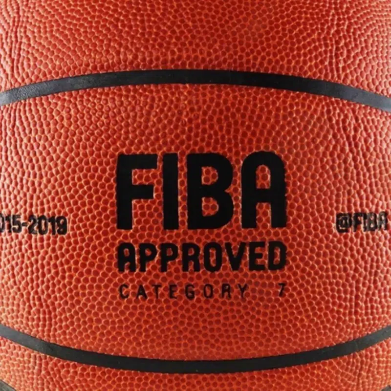 Primer plano de un balón de baloncesto naranja con el texto negro "FIBA Approved Category 7" (Categoría 7 aprobada por la FIBA). La superficie texturizada muestra los detalles de un Balón de Básquetbol MIKASA BQ1000 N°7, con los números "2015-2019" y la inscripción "(at)fiba".