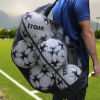 Una persona con una camisa azul lleva un Bolso Portabalones Tren lleno de pelotas de fútbol blancas y negras a través del campo de fútbol verde, donde líneas blancas nítidas contrastan con las montañas y árboles borrosos en el fondo.