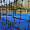 Una vista en primer plano de una red negra en una cancha de tenis al aire libre de color azul capta la atención. En el fondo, una pelota Red Paddle Uk Time Sport de 10 m x 0,92 m reposa sobre el césped entre árboles ligeramente borrosos y un cielo despejado, lo que sugiere que se aproxima un partido.