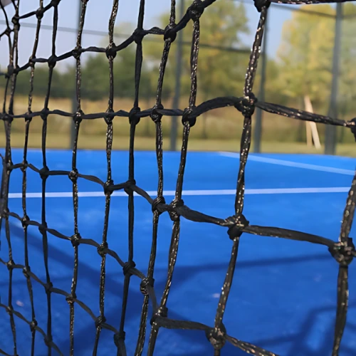 Una vista en primer plano de una red negra en una cancha de tenis al aire libre de color azul capta la atención. En el fondo, una pelota Red Paddle Uk Time Sport de 10 m x 0,92 m reposa sobre el césped entre árboles ligeramente borrosos y un cielo despejado, lo que sugiere que se aproxima un partido.